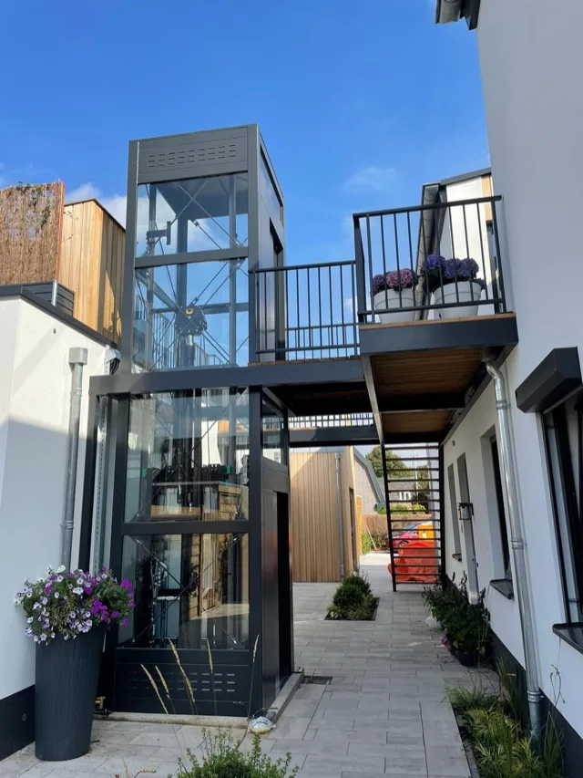 Black-framed outdoor glass elevator serving a terrace with flower pots