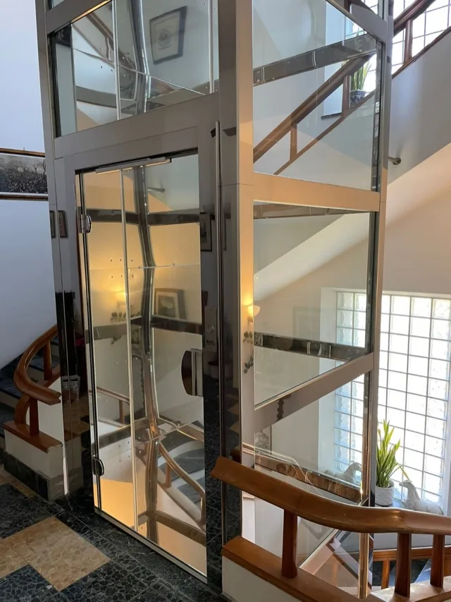 Glass and polished steel elevator beside a staircase with a wooden handrail