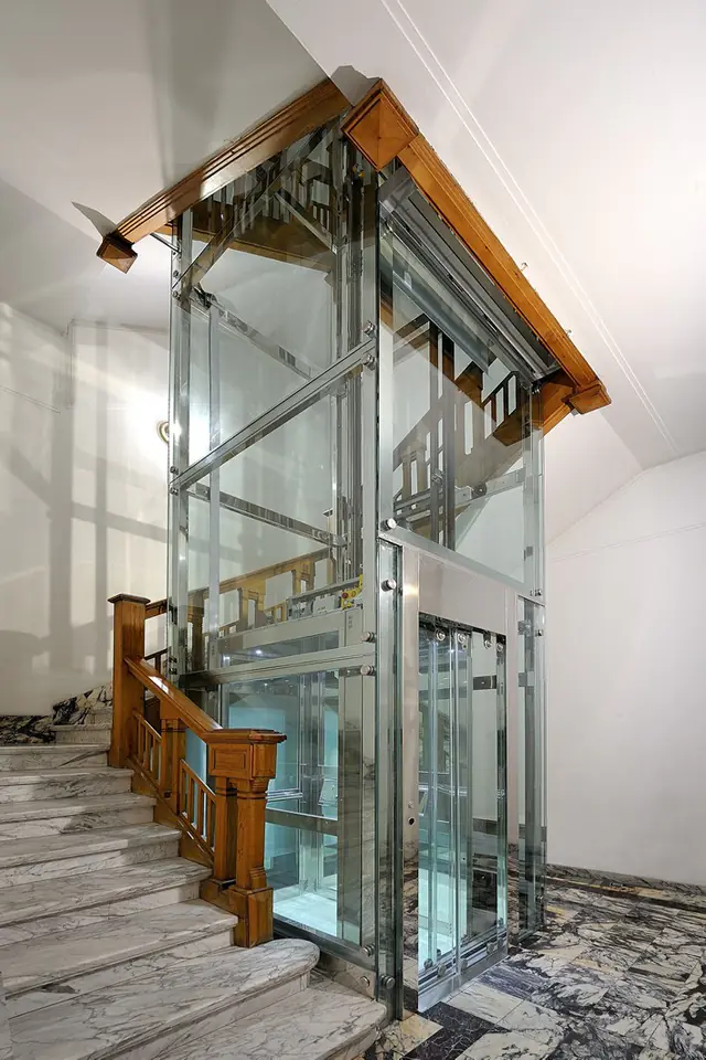 Glass elevator structure beside a marble staircase with a wooden handrail
